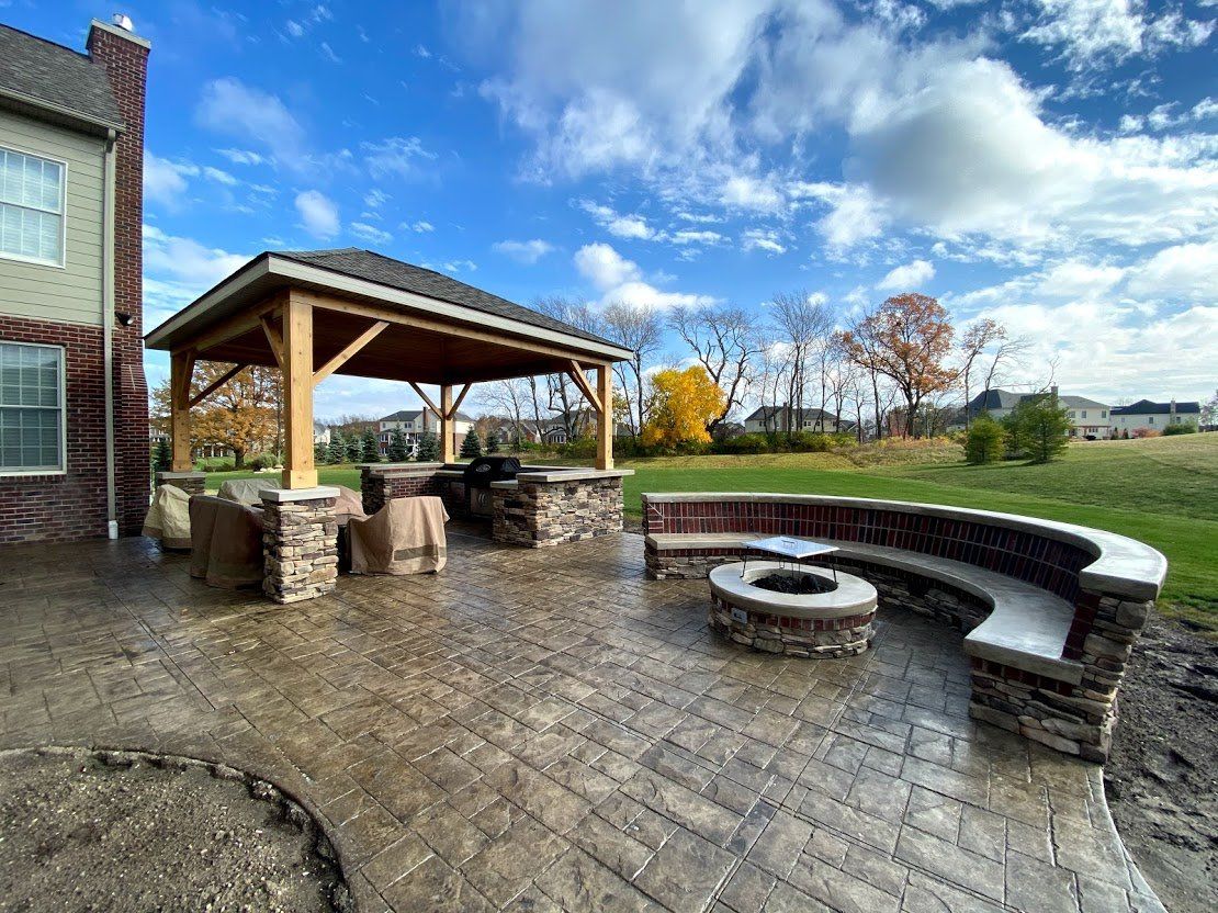 Stone patio with a covered pavilion, curved bench, and fire pit under a cloudy blue sky