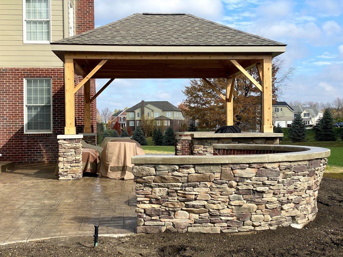 Stone gazebo with a shingled roof beside a building, overlooking a landscaped neighborhood.