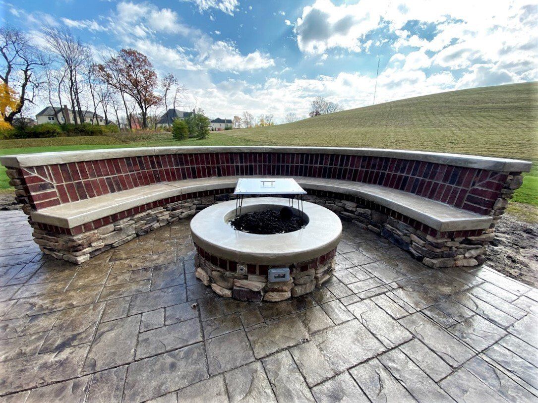 Stone fire pit with curved benches on a patio, overlooking a grassy hill and cloudy sky.