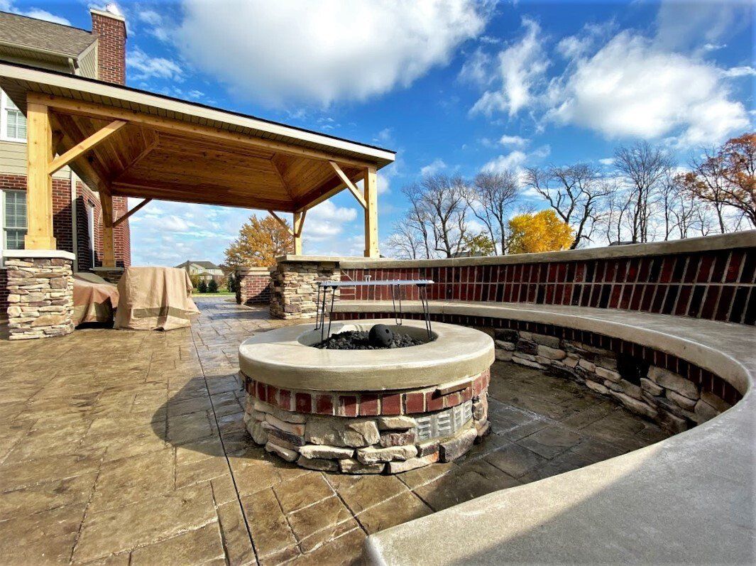 Outdoor stone fire pit with curved bench under a wooden pavilion on a sunny fall day
