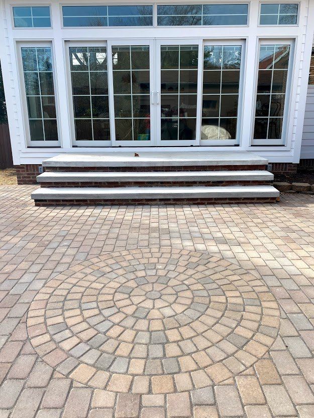 Brick patio with a circular pattern leading to white-framed glass doors and steps on a house exterior