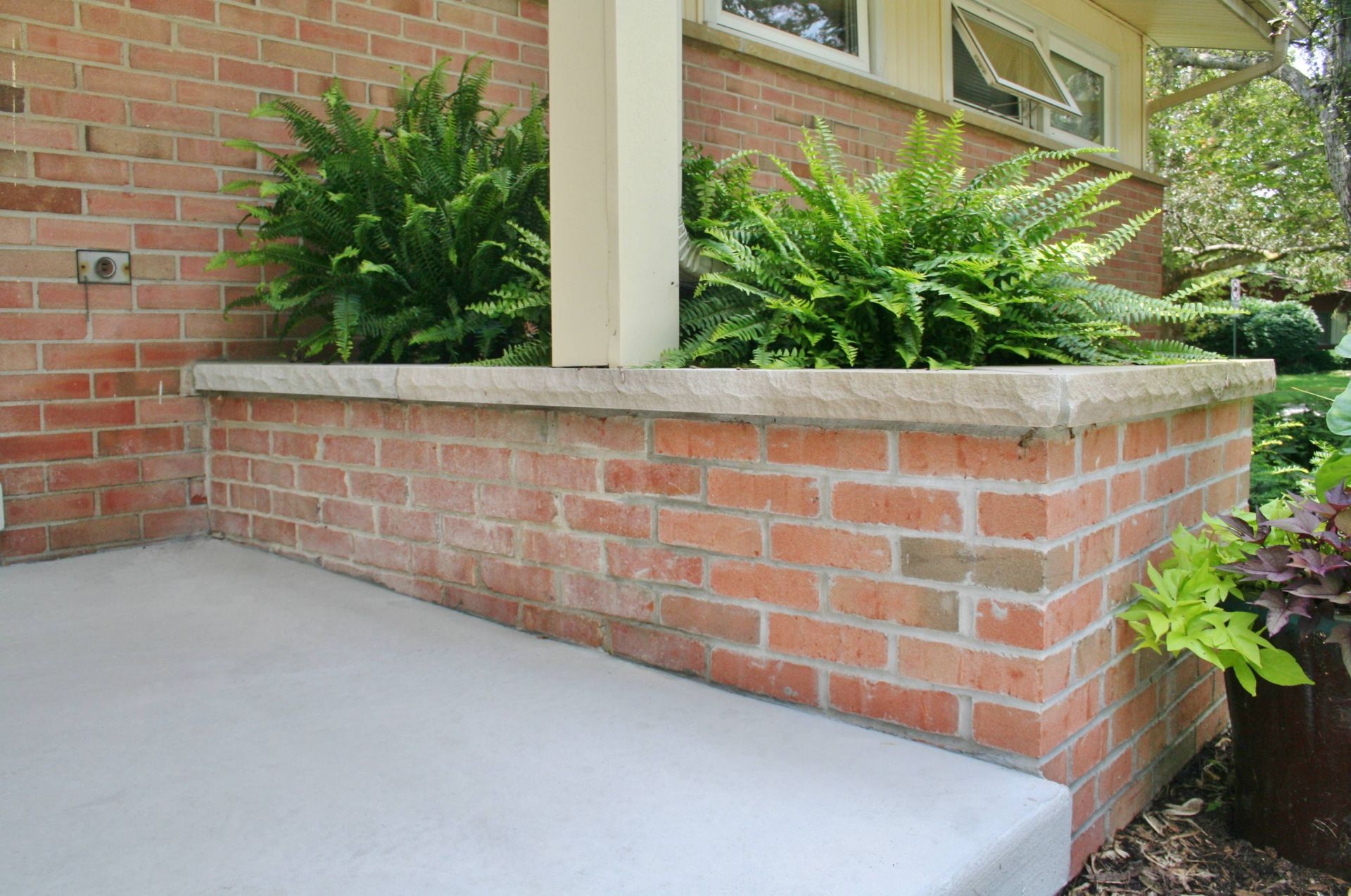 Brick planter beside a concrete patio with green shrubs in front of a brick house
