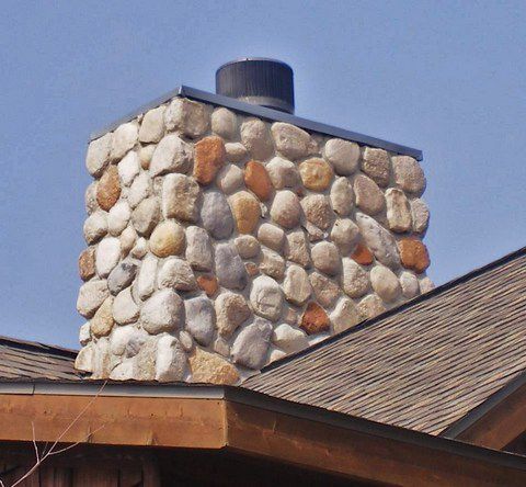 Stone chimney with a black cap rising from a shingled roof under a blue sky