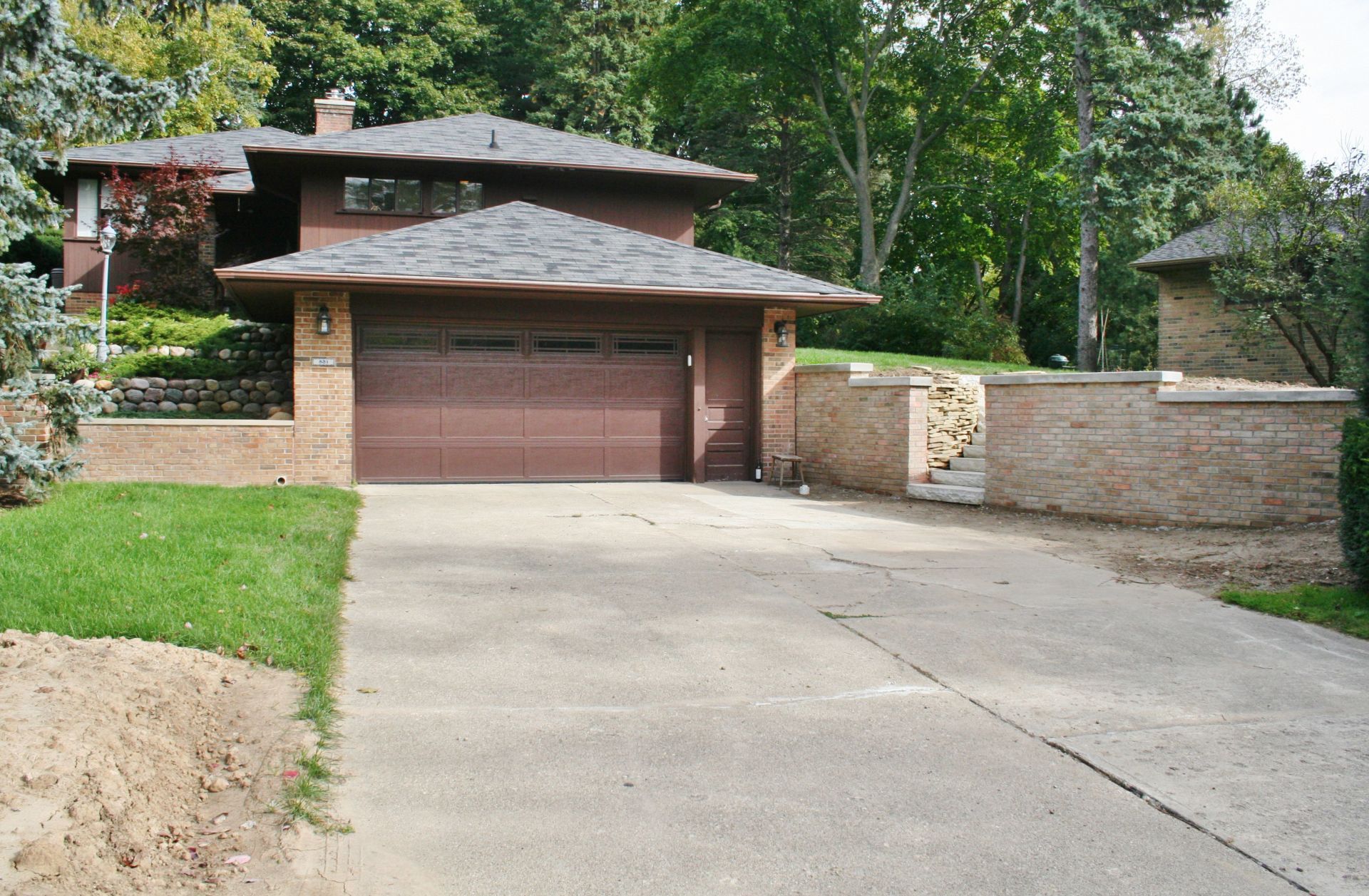 Brick house with brown garage door at the end of a concrete driveway, surrounded by trees and a stone wall