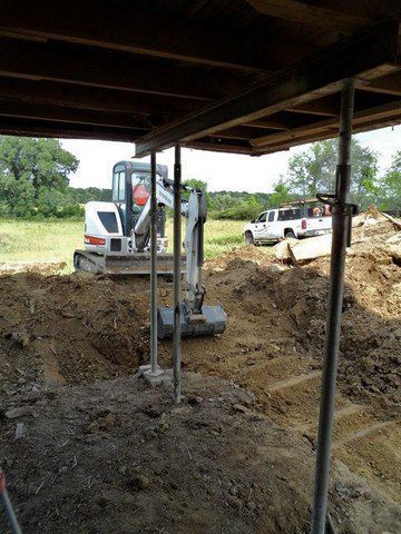 Construction site with excavator digging soil beneath a covered structure, trucks in the background