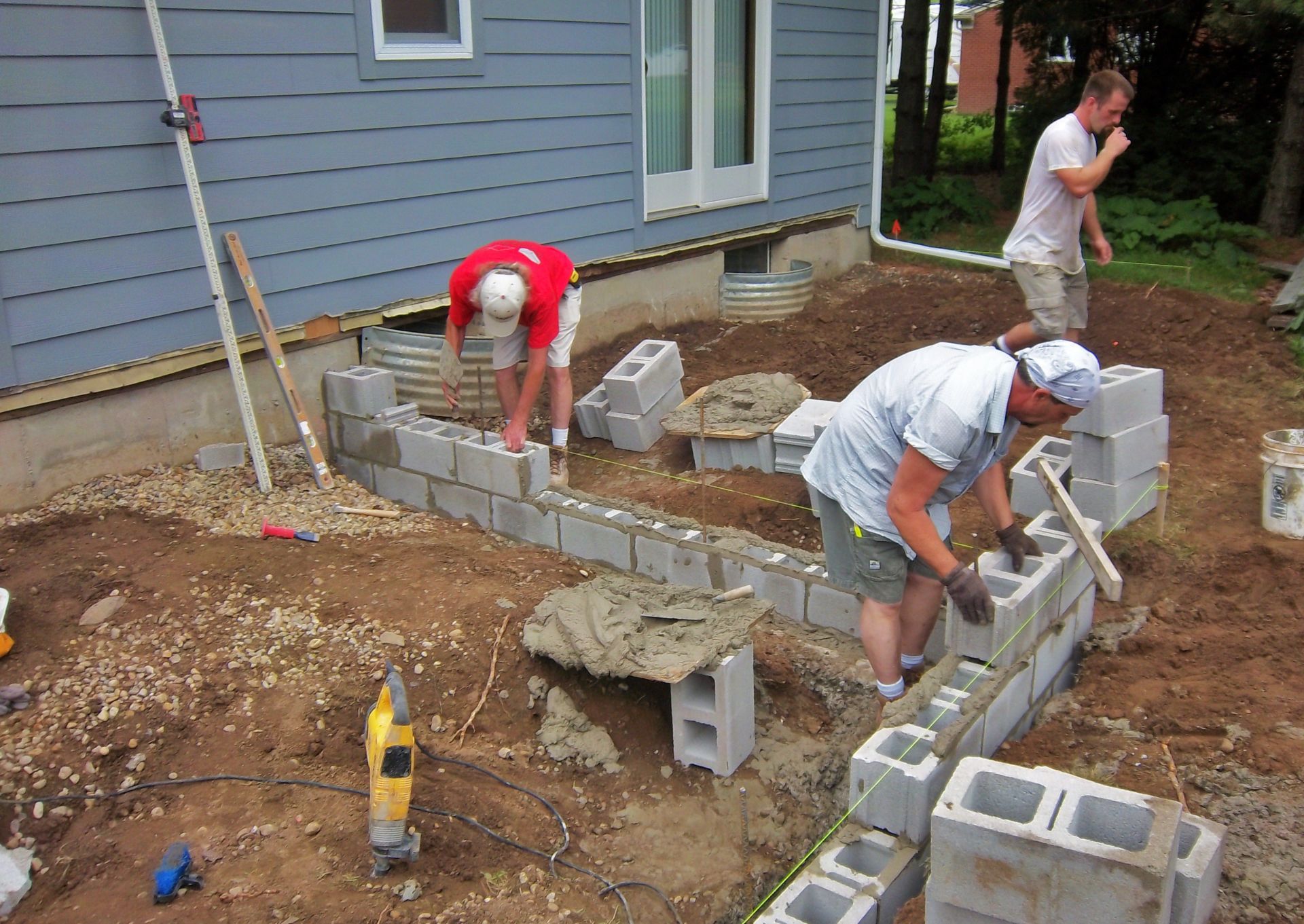 Workers laying cinder blocks to build a foundation beside a house.