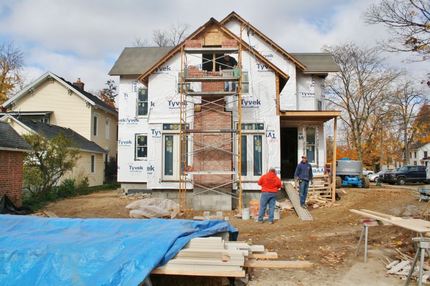 Two-story home addition under construction with brick chimney being built and house wrapped in Tyvek