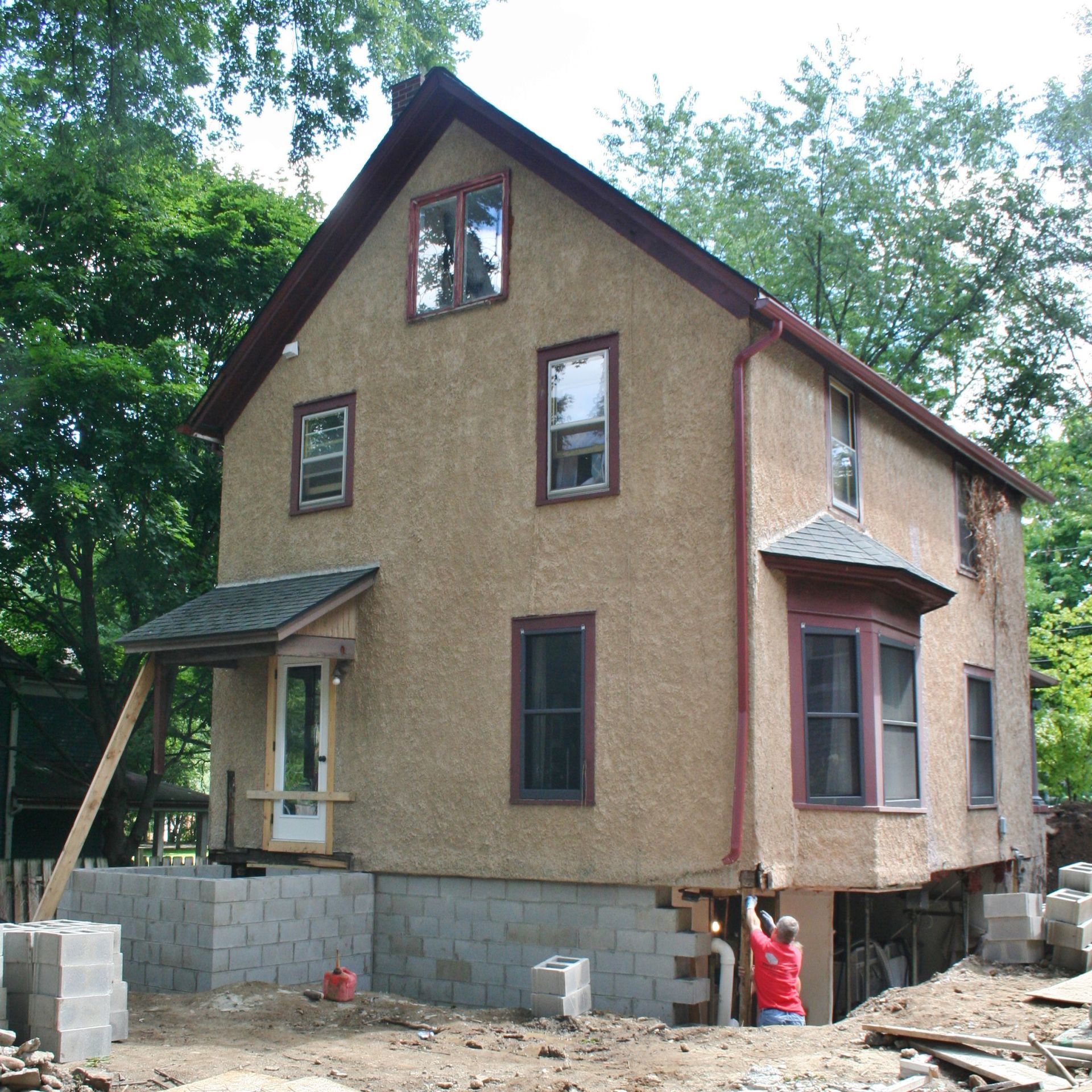 Partially built tan house with a brown roof, construction materials, and people working outside near trees