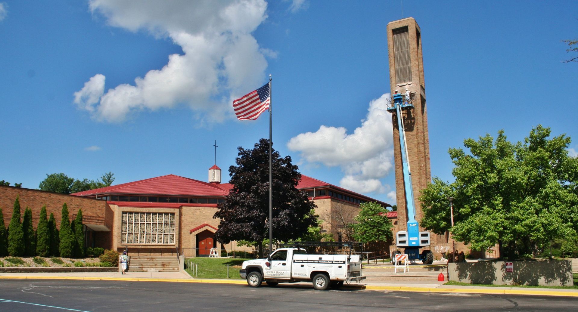 Brick building with red roof, tall bell tower, American flag, and a white truck parked outside under blue sky