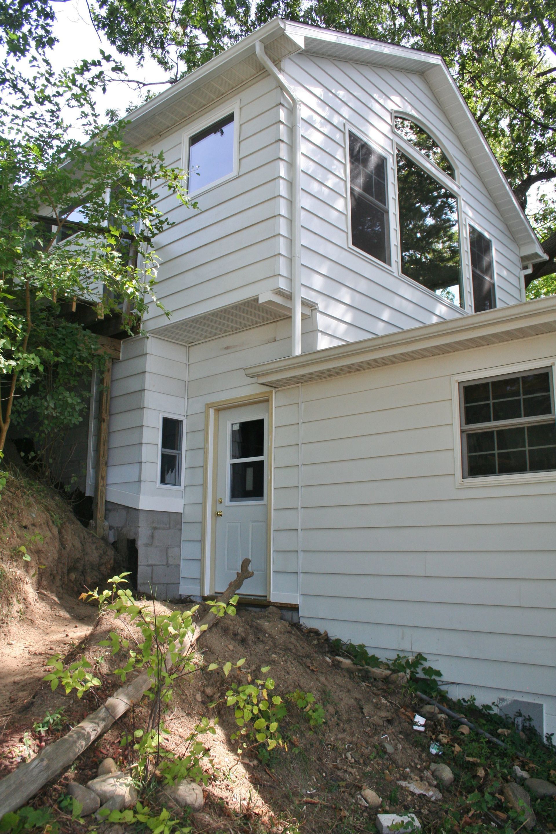 White house with a steep roof on a wooded hillside, with dirt and rocks in the foreground.