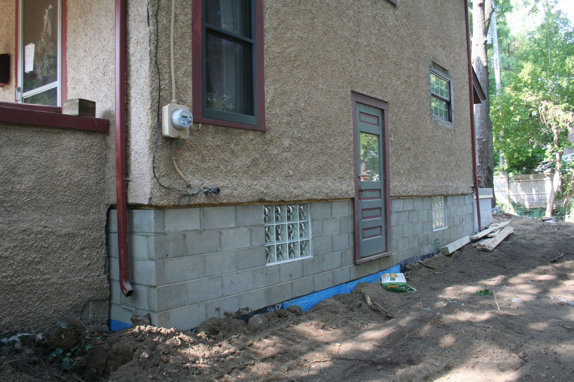Side of a house with a cinder block foundation, narrow door, and dirt yard with blue pipe along the wall