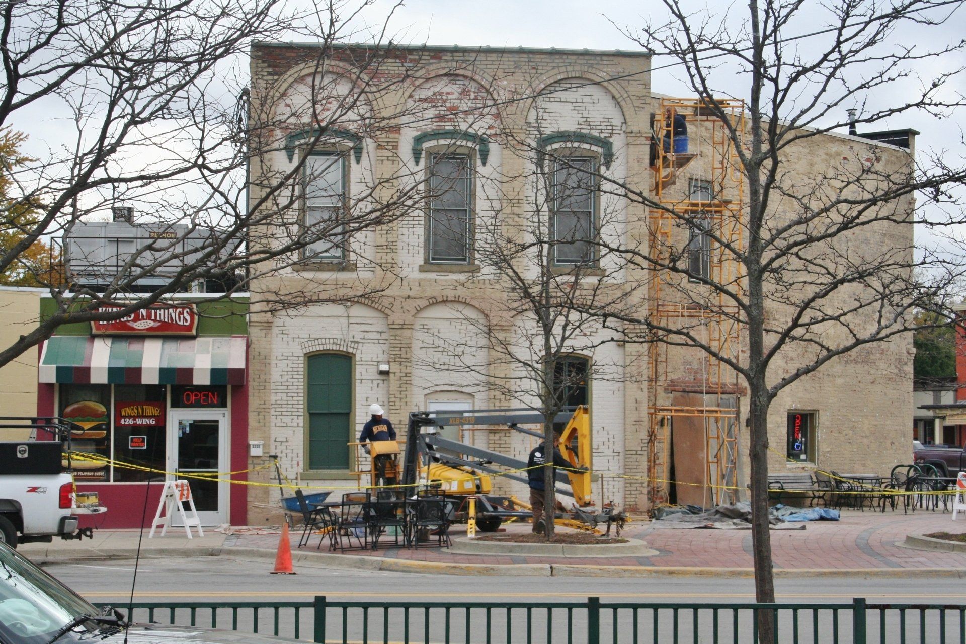 Historic stone building with arched windows and storefronts, outdoor café seating, and bare trees in front