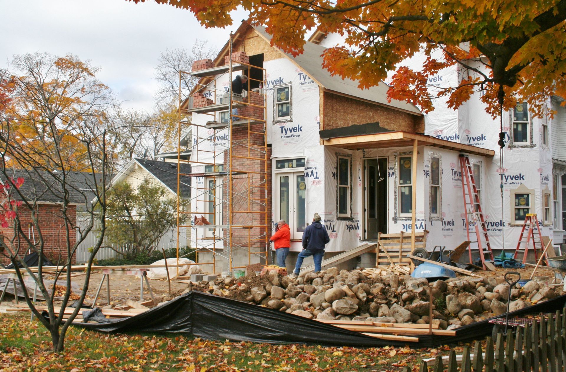 House under construction with workers, exposed framing, and rubble in front, framed by autumn trees