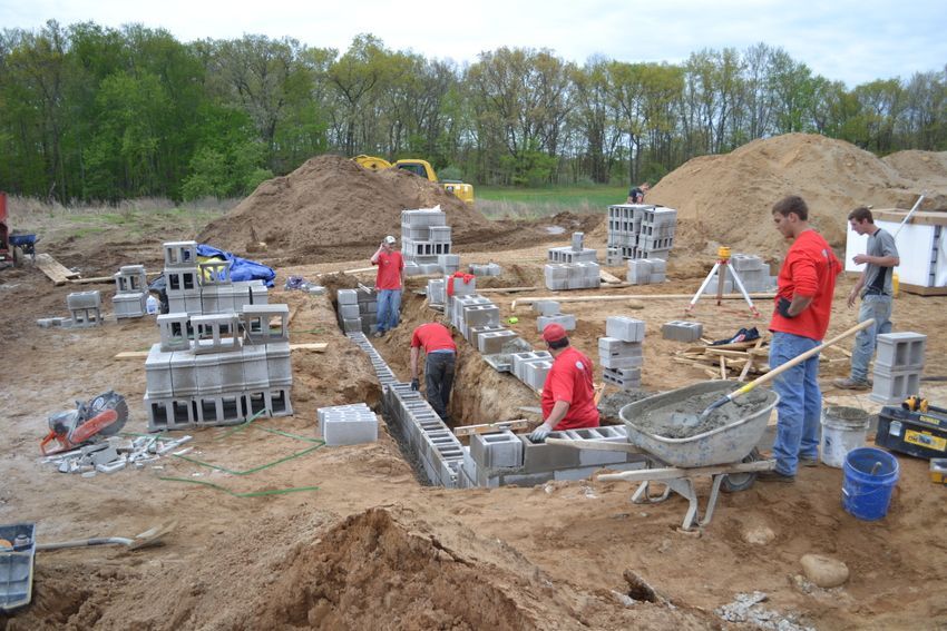 Workers laying cinder blocks at a construction site with wheelbarrows, tools, and dirt piles.