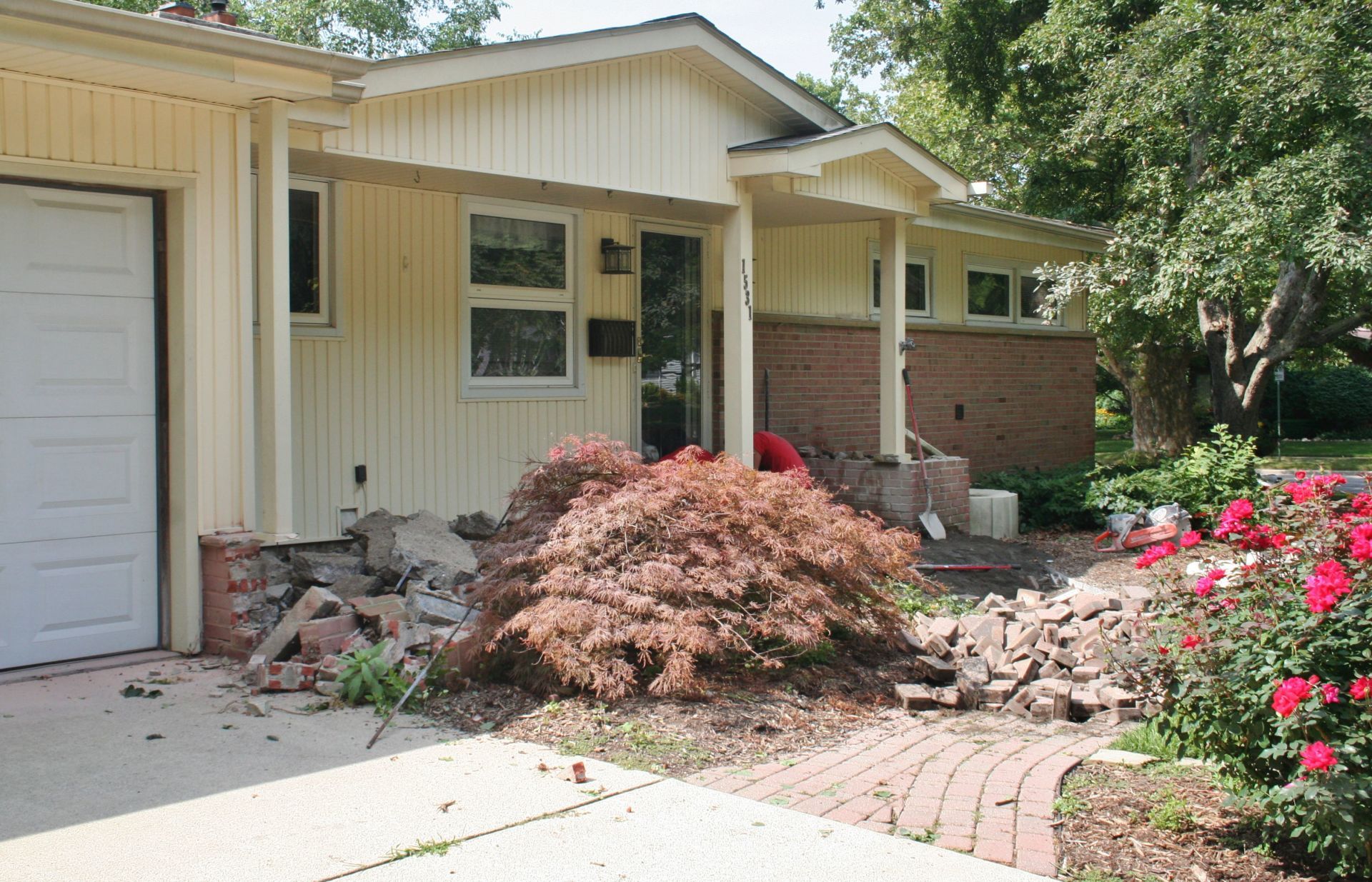 Front entrance of a house with a brick walkway, bushes, and a pile of debris near the garage