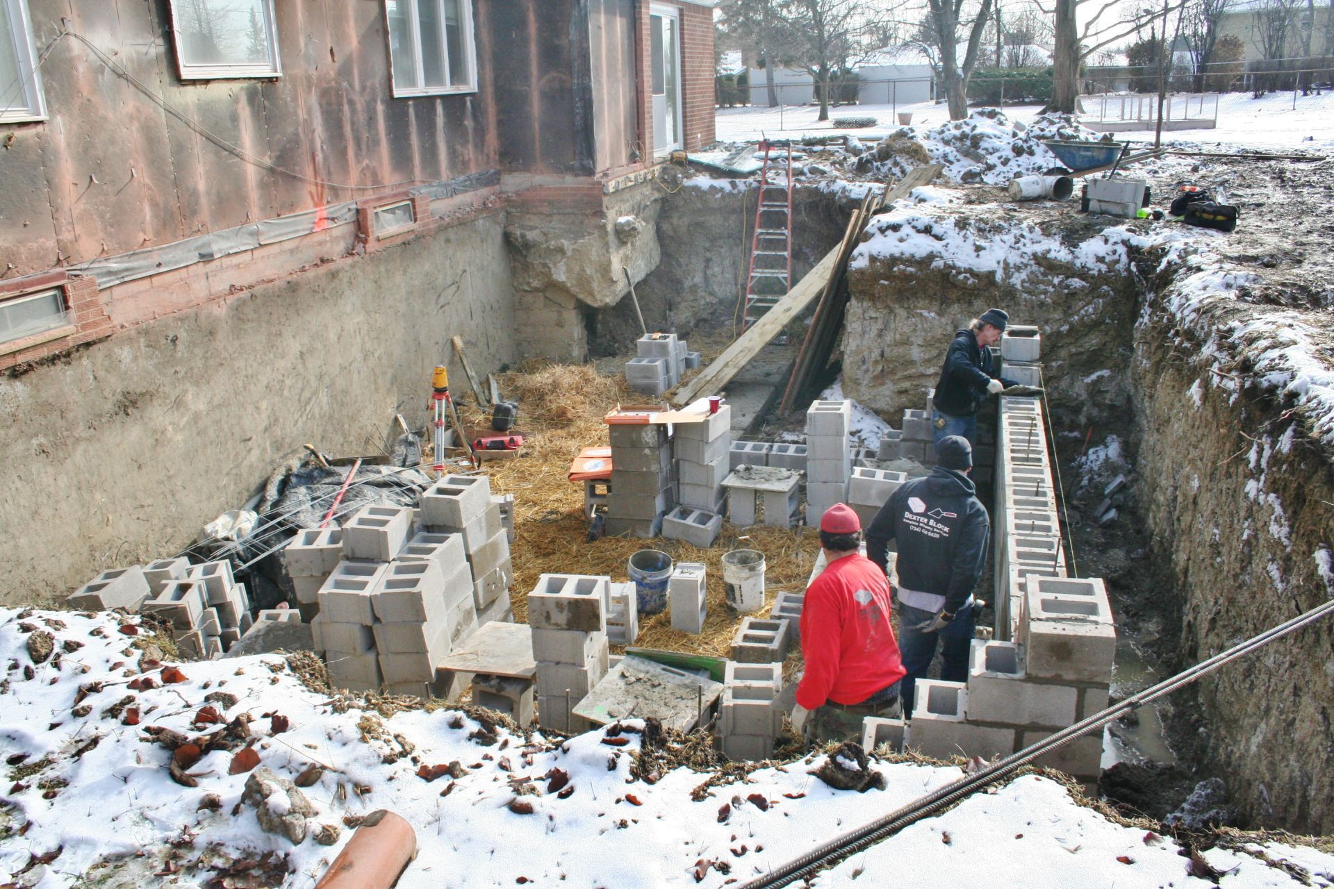Snowy construction site beside building, with workers laying cinder blocks in a deep trench.