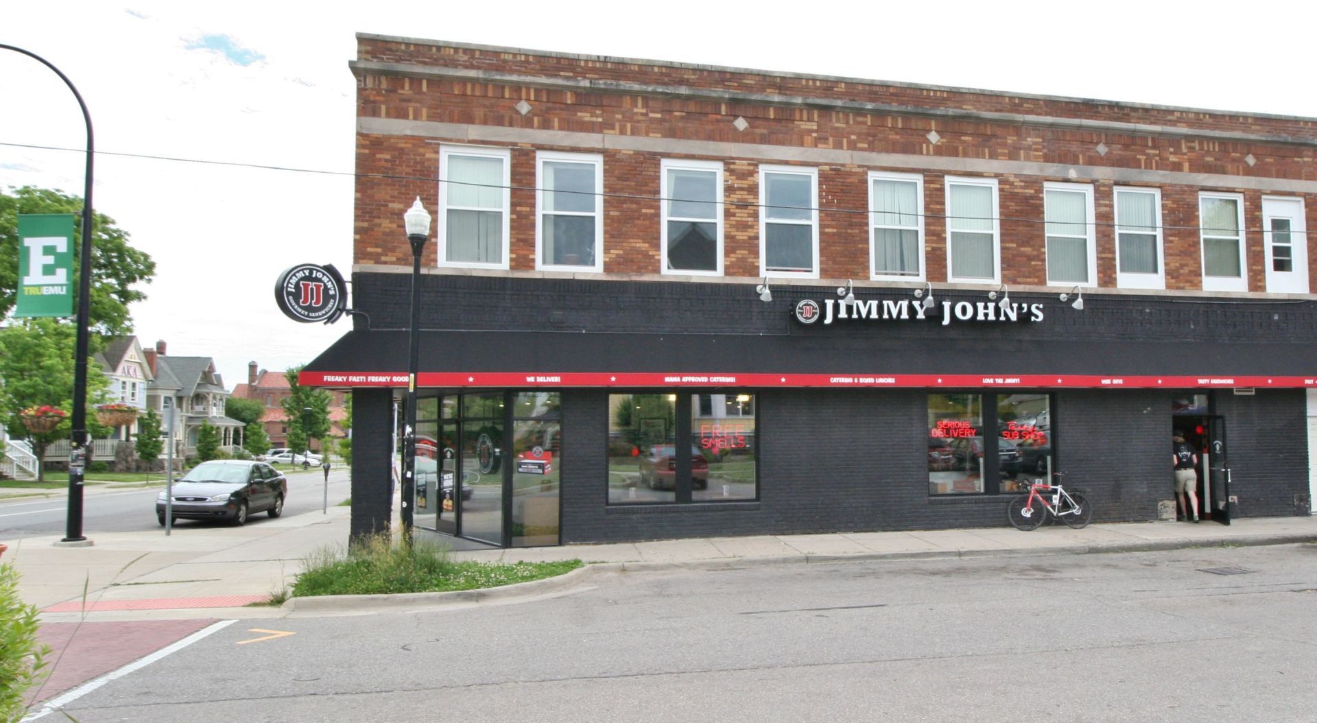 Street view of Jimmy John’s brick storefront with black awning and red trim on a city corner.