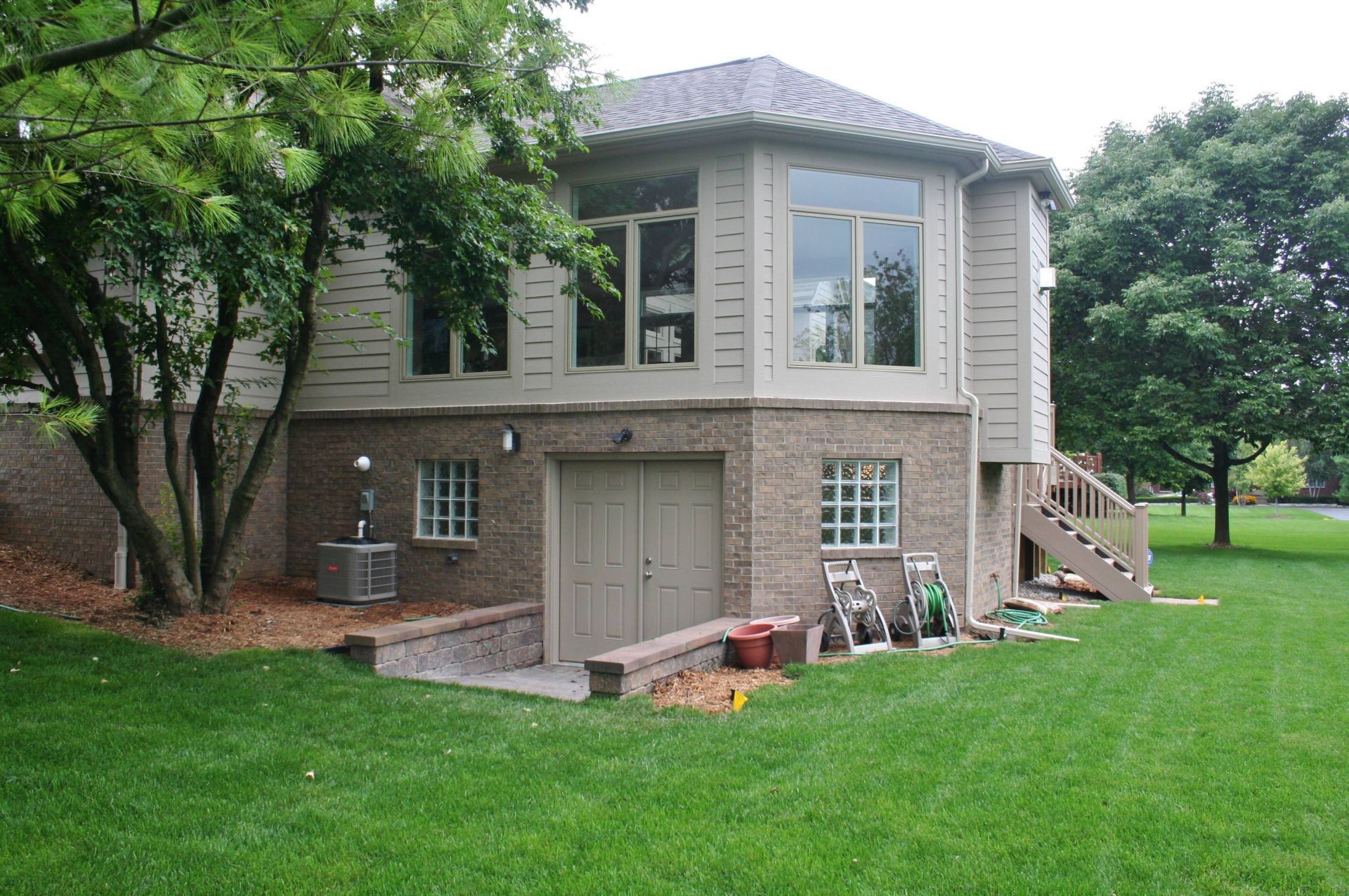 Two-story beige house with a large lawn, trees, and a garage beneath the main floor.