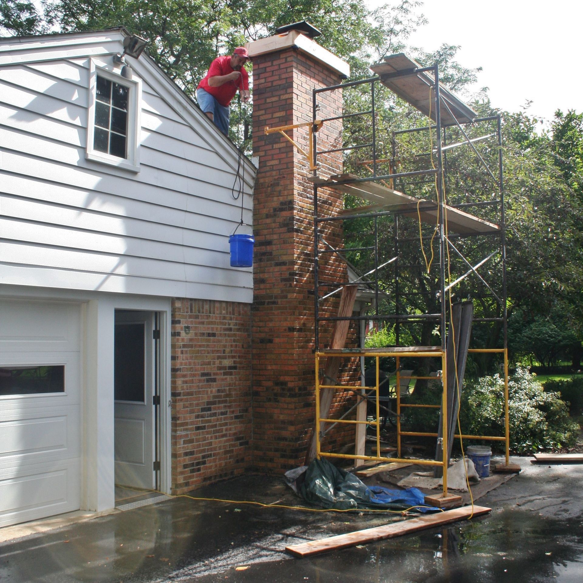 Worker on scaffolding repairing a brick chimney beside a house with an open garage.