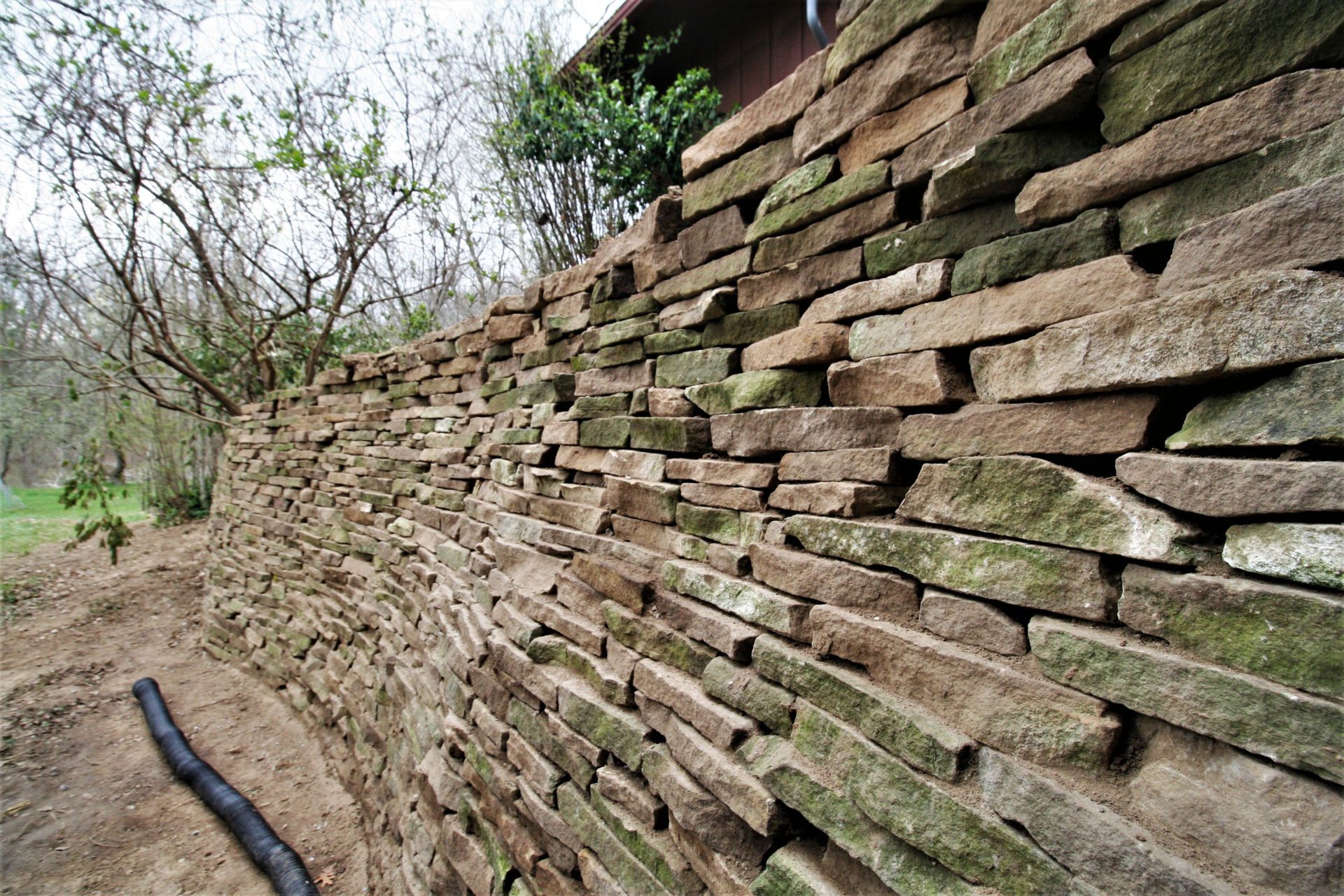 Stacked stone wall along a dirt path, with trees and greenery beside it.