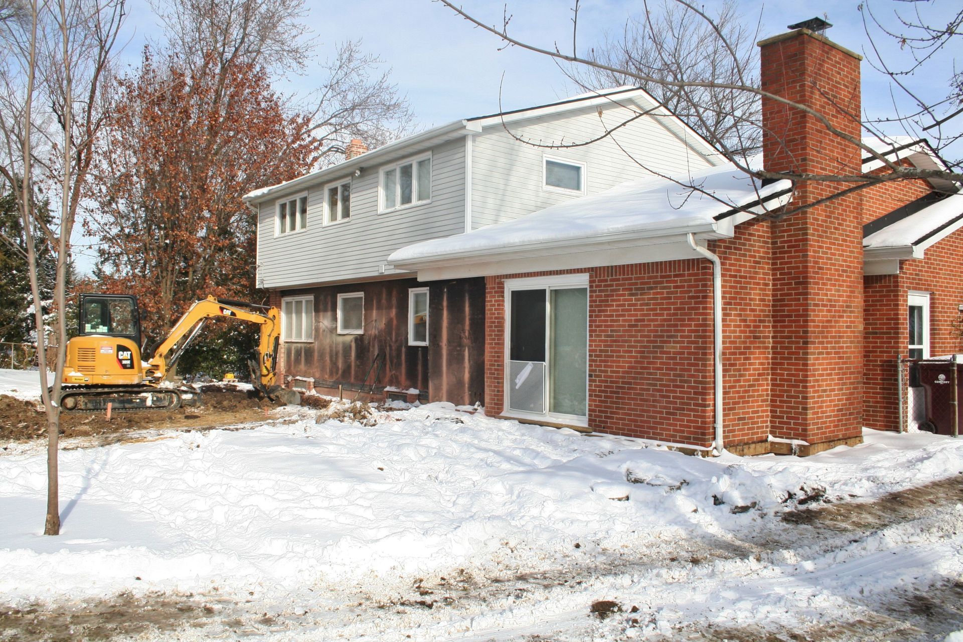 Snow-covered house with red brick and white siding, and an excavator working in the yard.