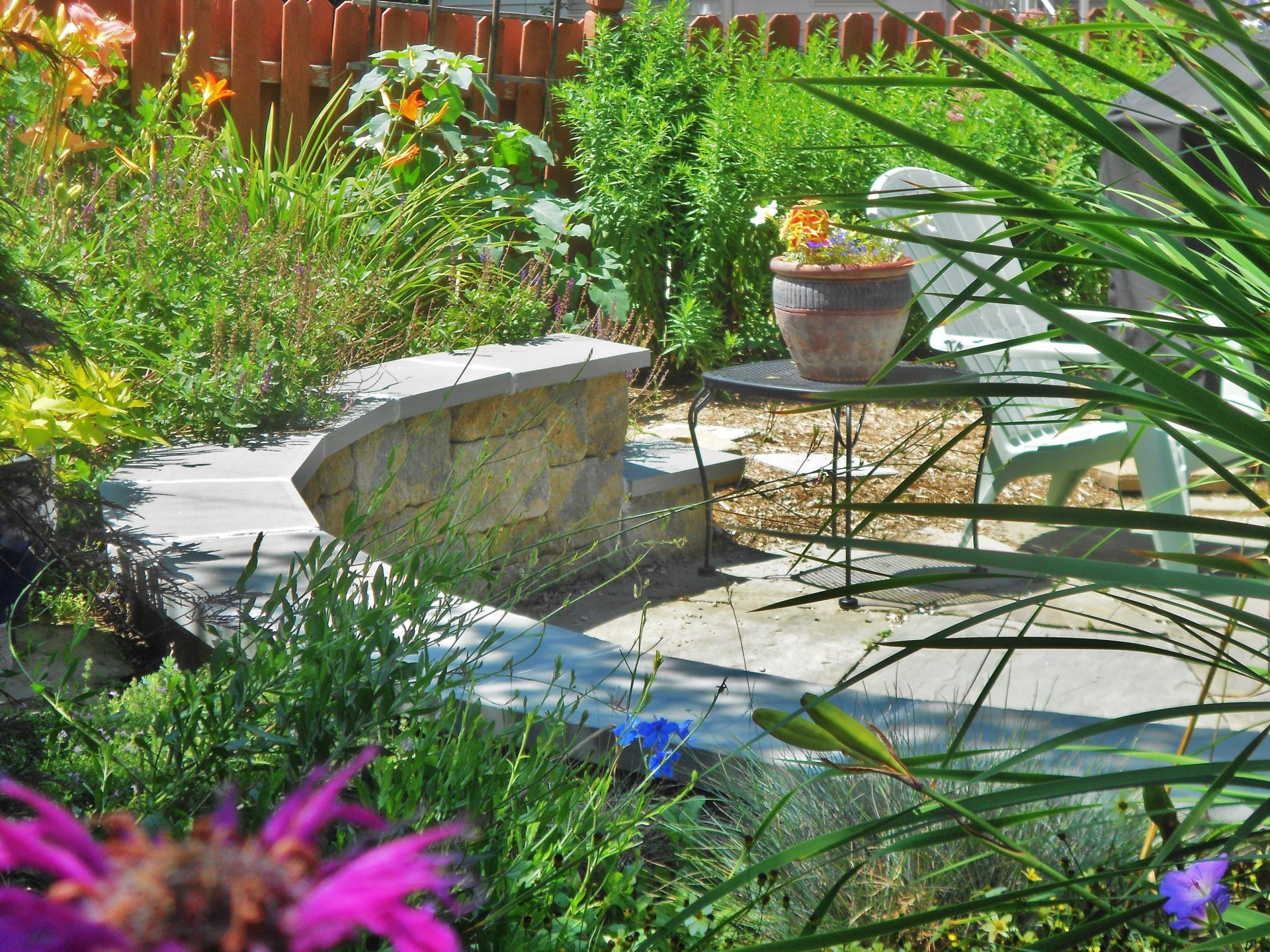 Sunlit garden patio with stone bench, metal table, and lush green plants surrounding a stone walkway