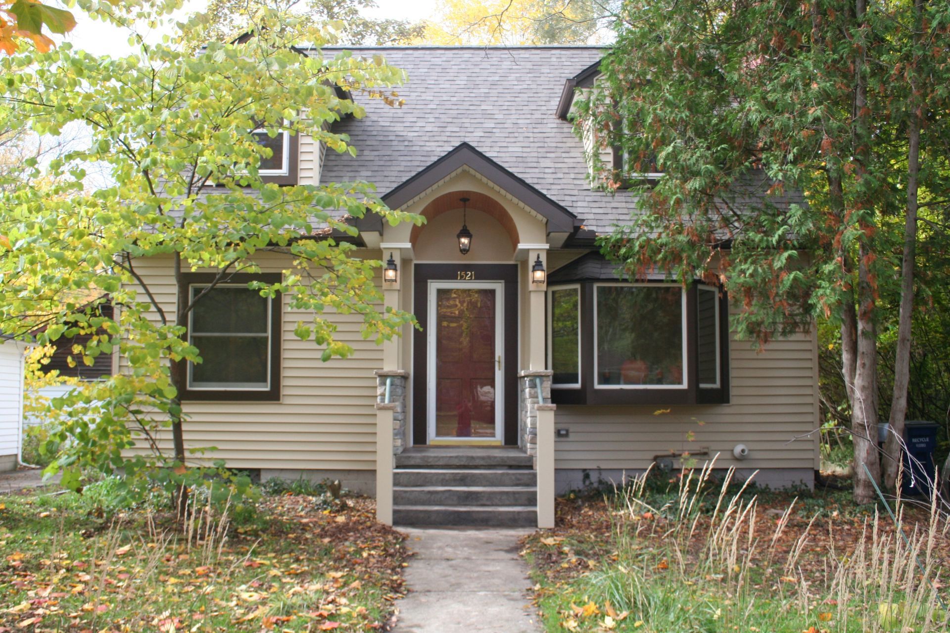 Small beige house with dark shutters, a front porch, and a tree-lined yard in autumn.