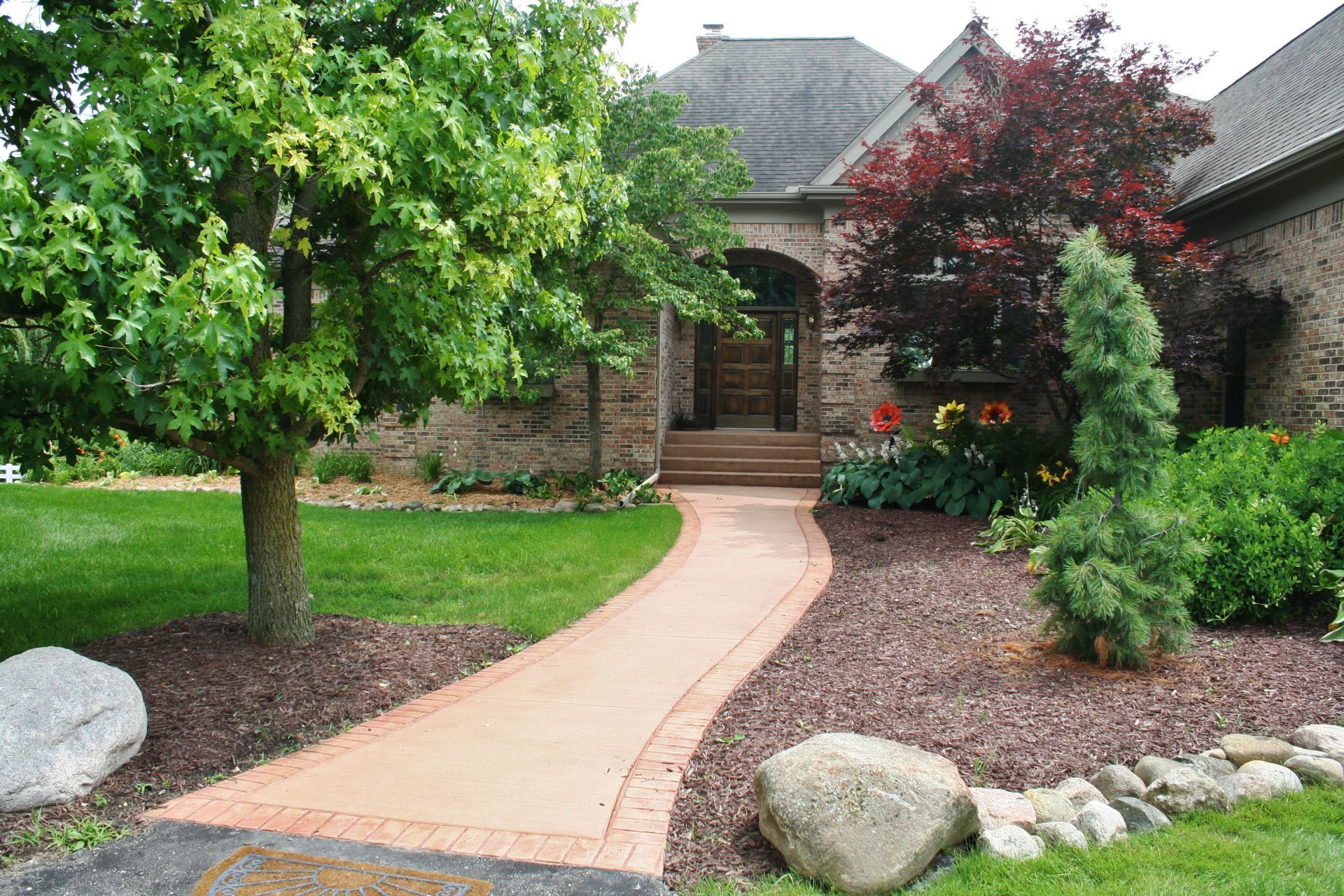 Curved walkway leading to a brick house entrance, framed by trees, shrubs, and landscaped flower beds.