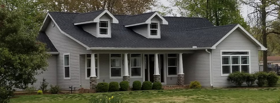 A light gray house with a dark shingled roof, front porch with columns, two dormer windows, and a lawn in front.