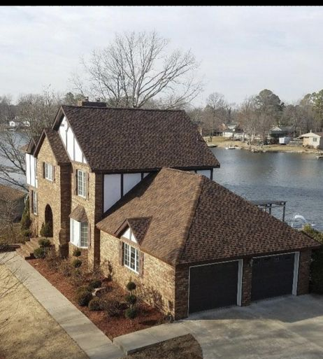 An elevated view of a brick Tudor-style home with a shingled roof, nestled on the shore of a calm lake.