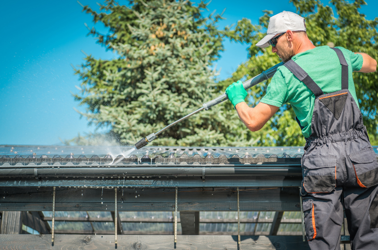 A worker in a green shirt and overalls power-washes a translucent corrugated roof against a blue sky.
