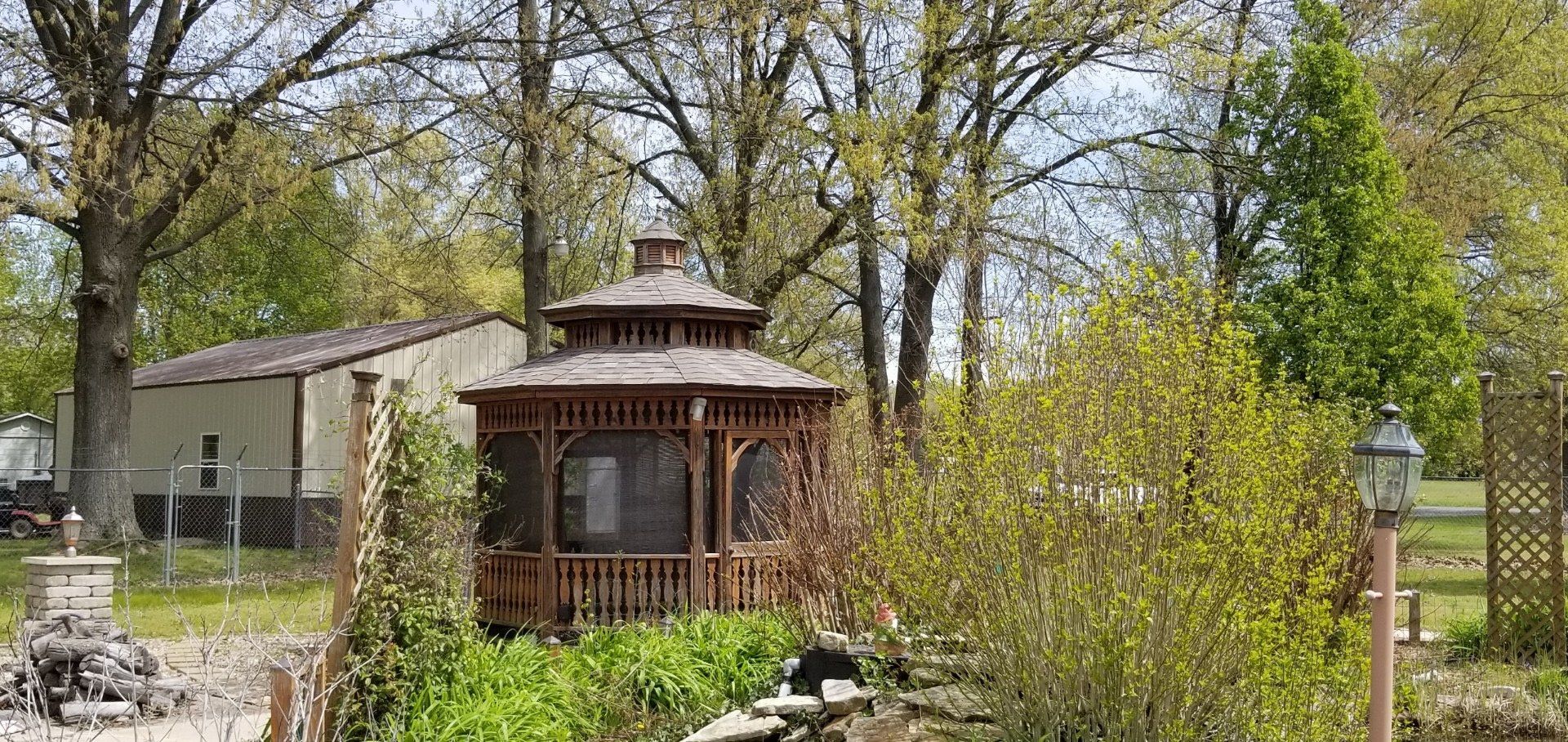 A wooden gazebo stands in a sunny, tree-filled garden with a small structure and fence in the background.