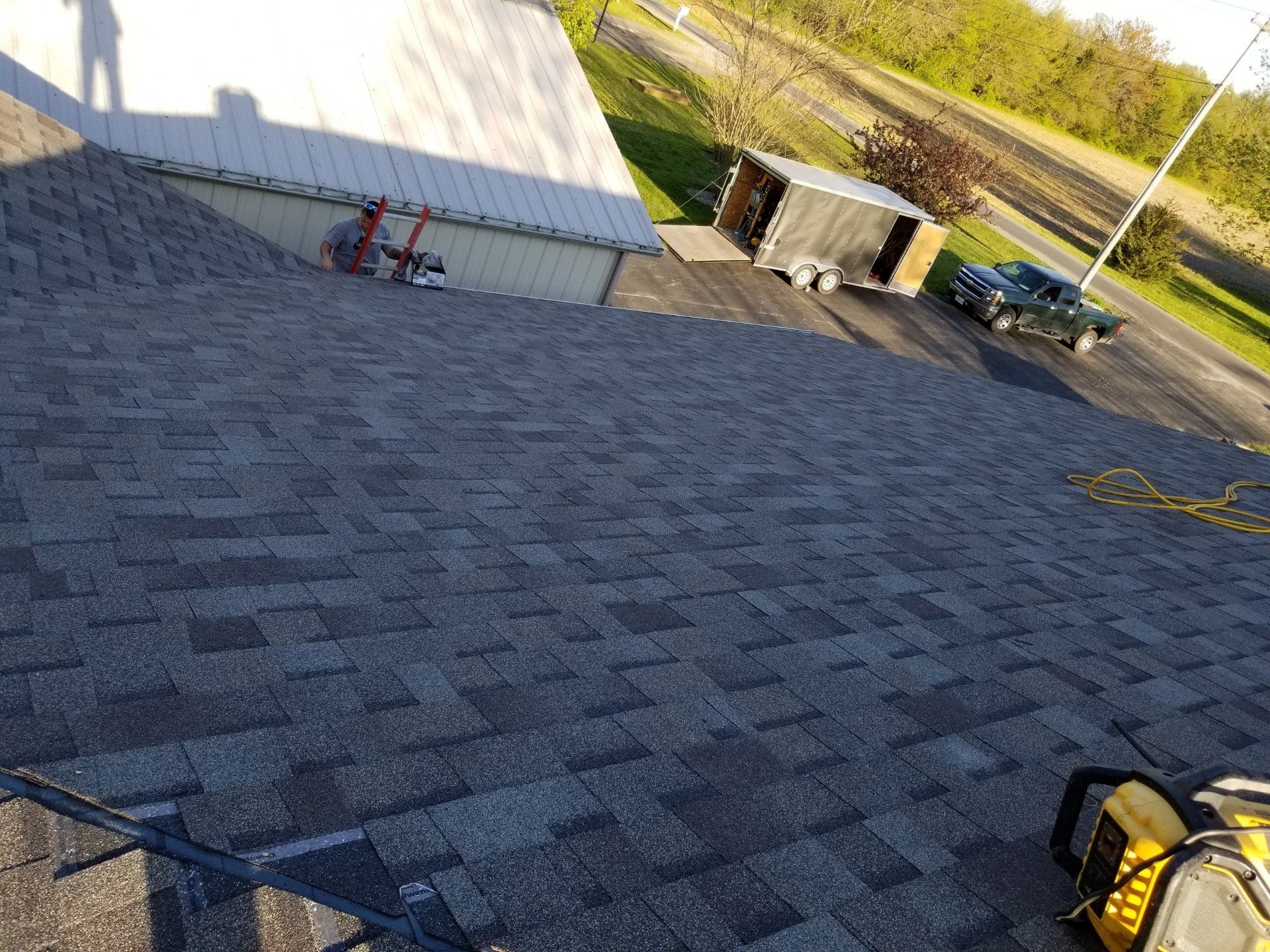 A view from a roof showing grey asphalt shingles in the foreground, with a trailer and a truck parked below.