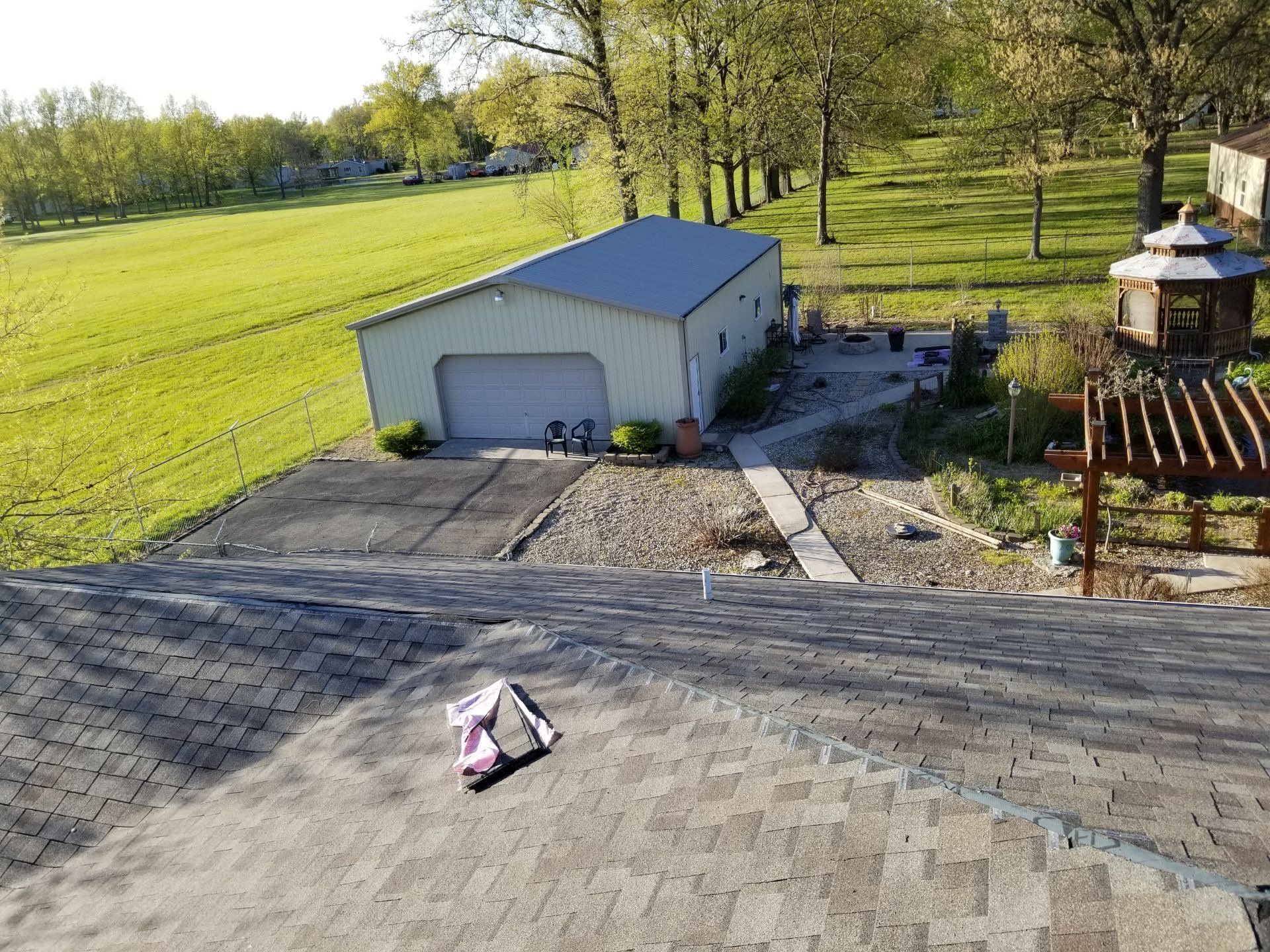 An aerial view from a shingled roof looking down at a light green detached garage, a gravel yard, and a wooden gazebo.