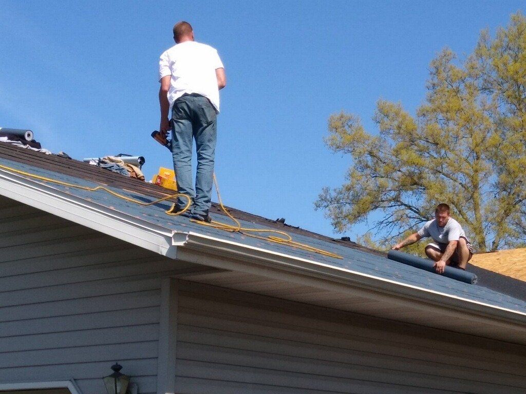 Two workers install roofing material on a slanted roof under a bright blue sky.