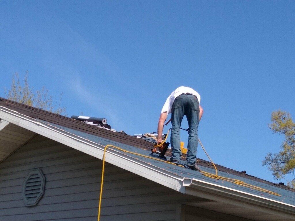 A person wearing jeans and a white shirt uses a nail gun to install dark asphalt shingles on a roof under a blue sky.