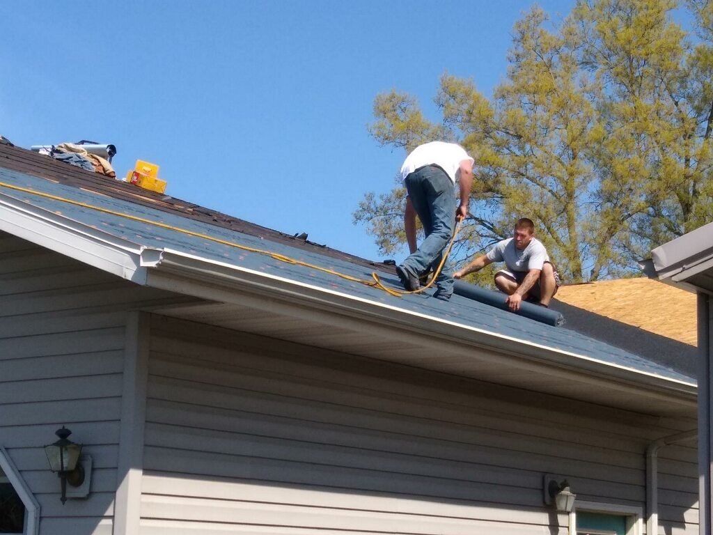 Two workers install gray roofing material on the sloped roof of a house under a clear blue sky.