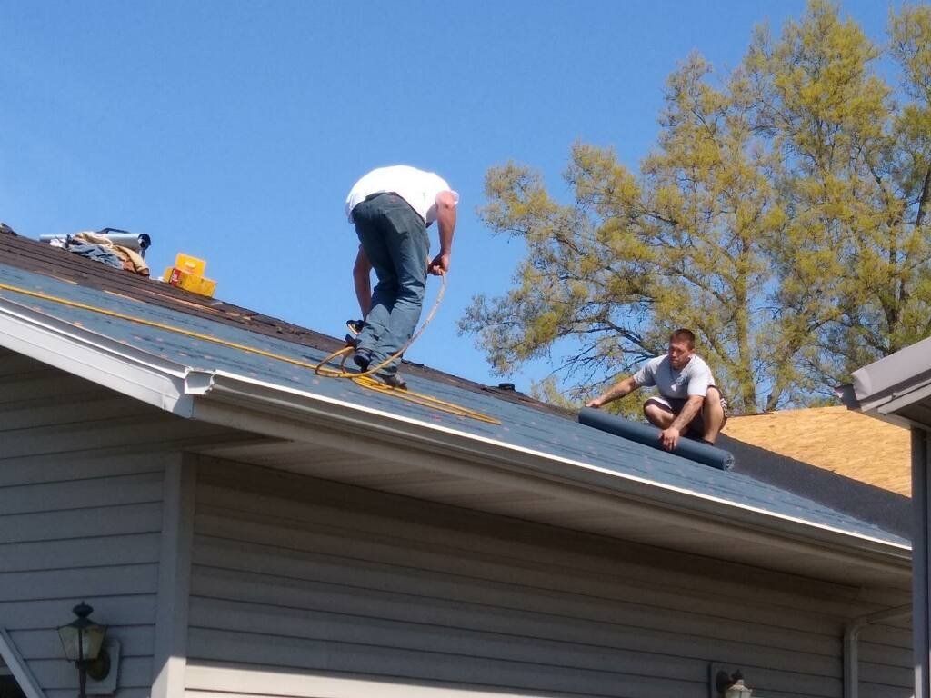 Two people working on a residential roof, installing black underlayment on a sunny day.
