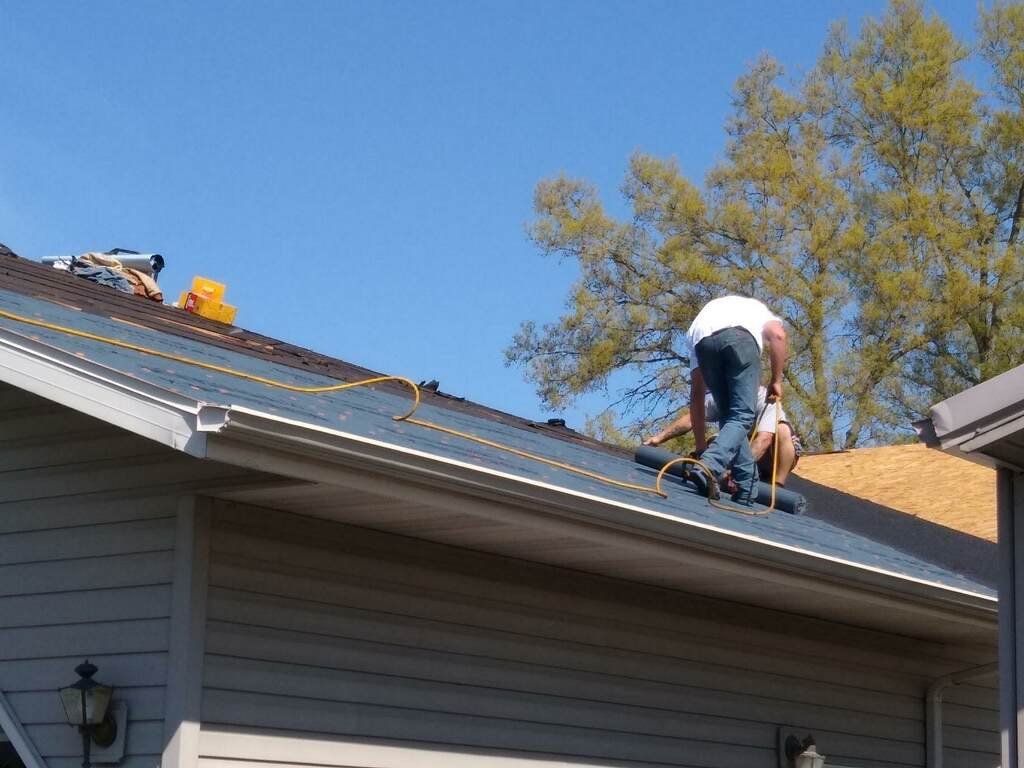 A worker in a white shirt and jeans kneels on a slanted, shingled roof under a clear blue sky.