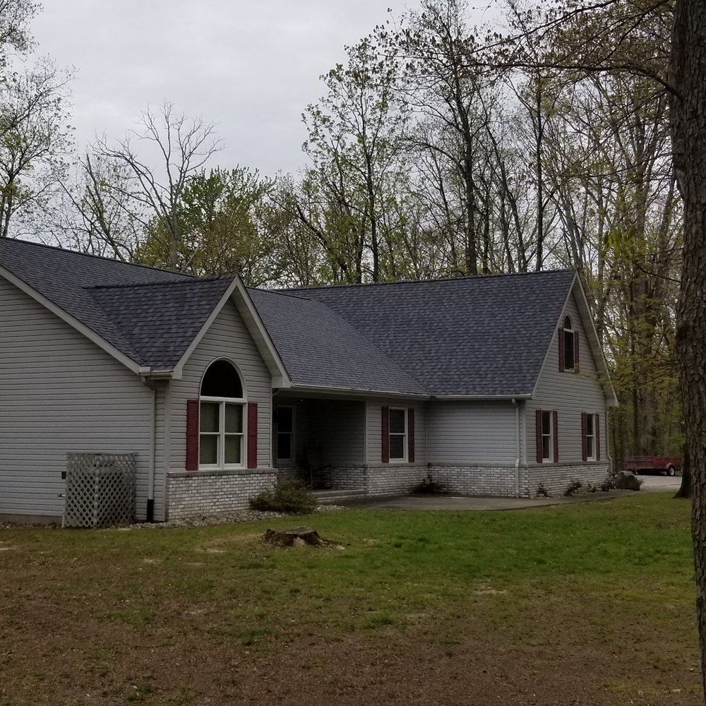 A gray, single-story house with a dark shingled roof, arched window, and stone foundation in a wooded yard.