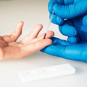 A una persona se le pincha el dedo con una pipeta para realizarle un análisis de sangre; se ve una mano enguantada de color azul y un kit de prueba.