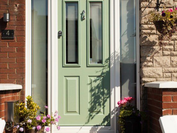 A green front door with flowers in front of it is on a brick house.