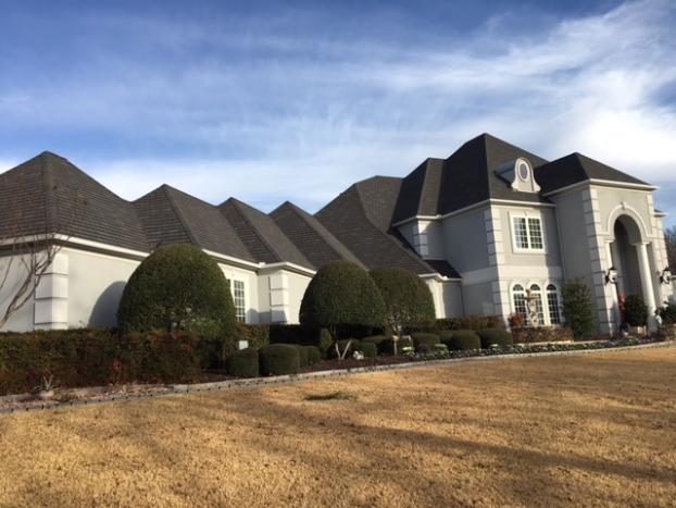 Large, light-grey house with multiple rooflines, oval window, and manicured shrubs, against a blue sky.