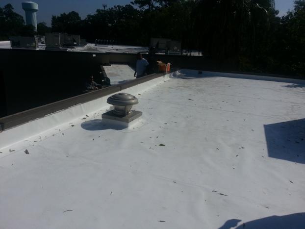 White commercial roof with vent; worker applying sealant. Blue sky, water tower in background.