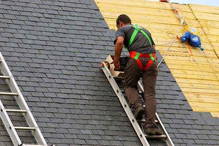 Roofer on a ladder with safety harness, installing dark gray roof tiles on a house.