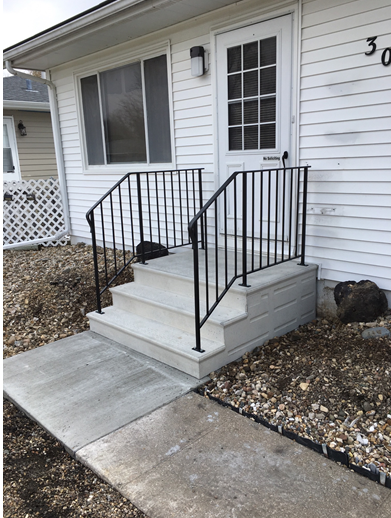Front porch steps with black railings leading to a white house entrance and concrete walkway