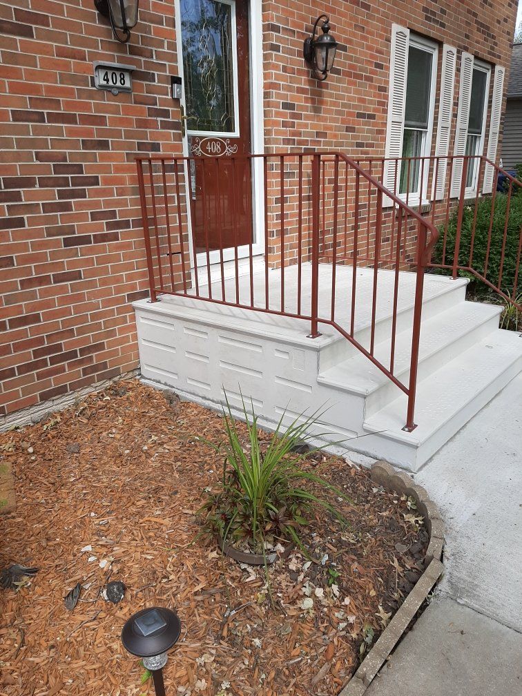 Brick house front stoop with white steps, red railing, and small garden bed.