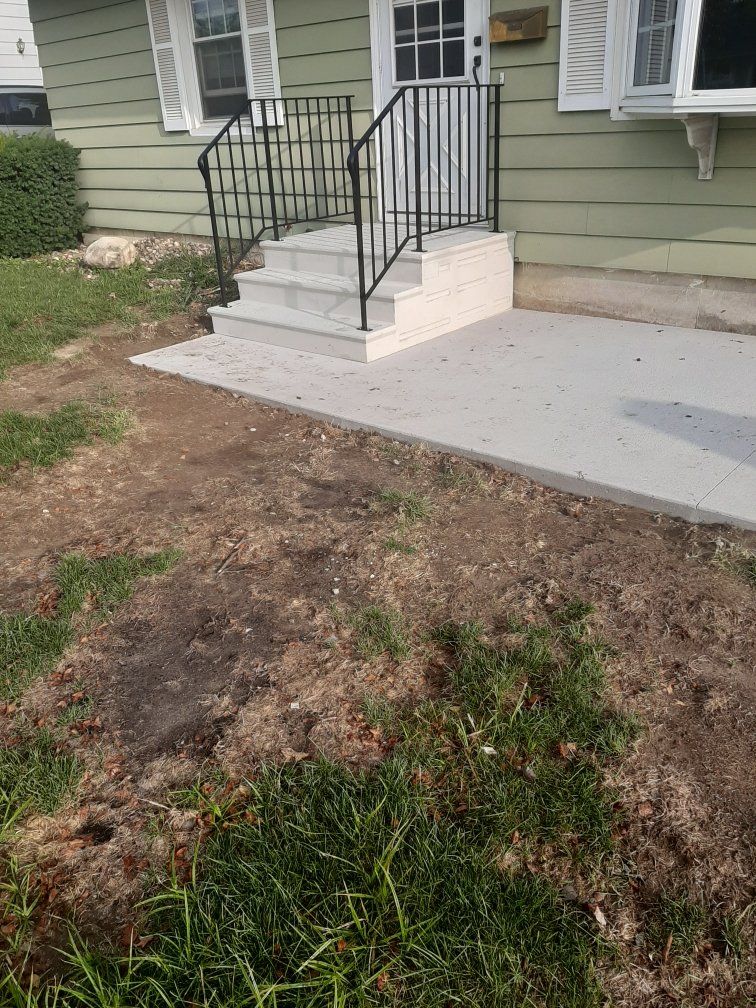 Concrete front steps with black railings leading to a green house, beside a bare dirt yard.