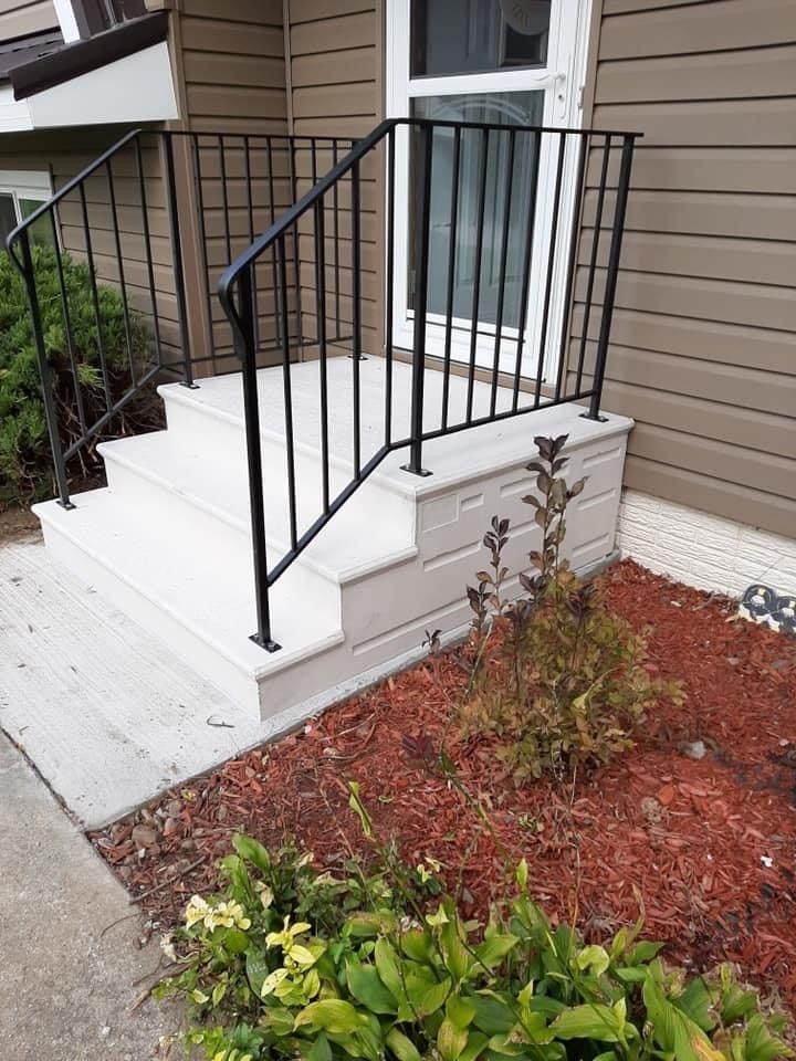 Front steps with black railing, white stoop, and brown siding beside a small red mulch garden.
