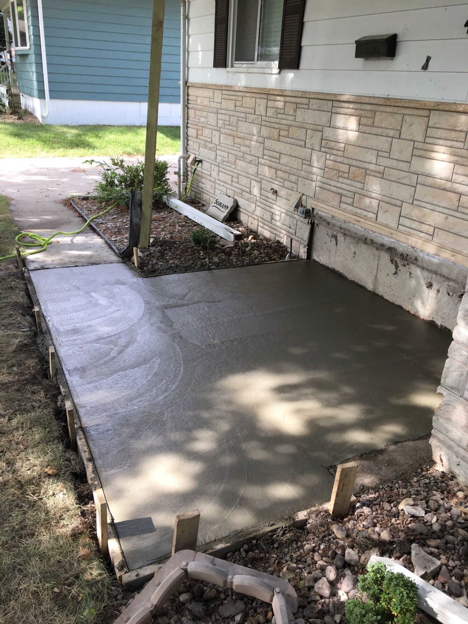 Fresh concrete patio slab beside a house, bordered by stakes and a brick foundation